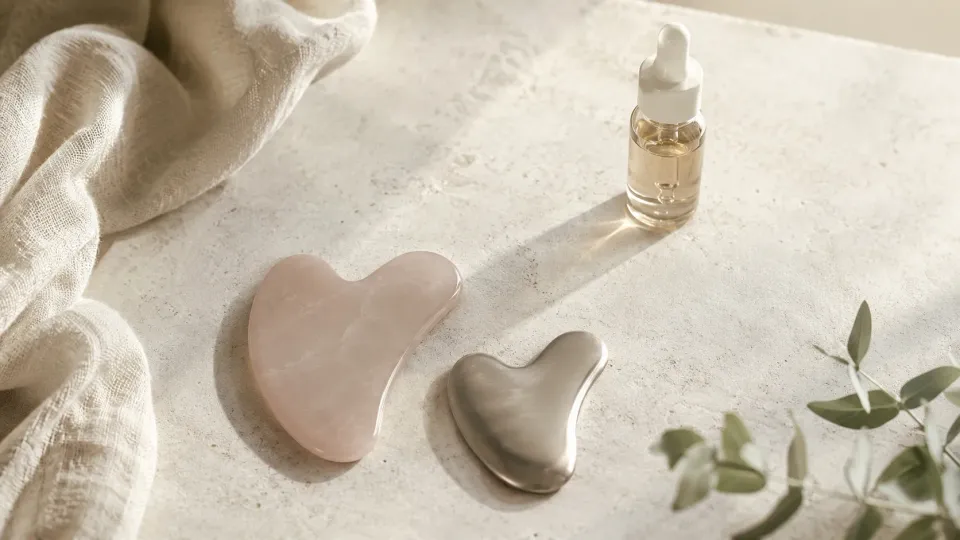 Deux gua sha, l’un en pierre rose et l’autre en métal, posés près d’un flacon de sérum.